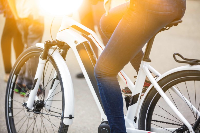 E-Bike Nahaufnahme eines E-Bikes, der Fokus liegt dabei auf der Batterie in der Radmitte. Auf dem Rad sitzt jemand, man sieht jedoch nur die Beine.