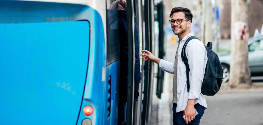 Ein junger dunkelhaariger Mann mit Brille steigt lächelnd in einen blauen Bus.