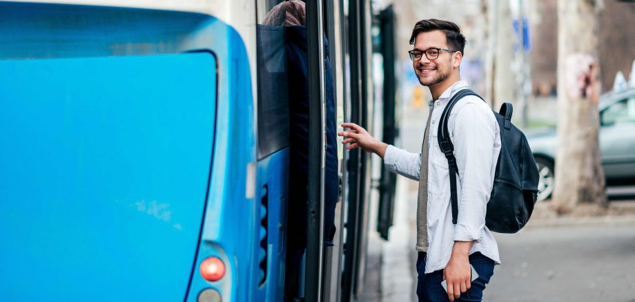 Ein junger dunkelhaariger Mann mit Brille steigt lächelnd in einen blauen Bus.