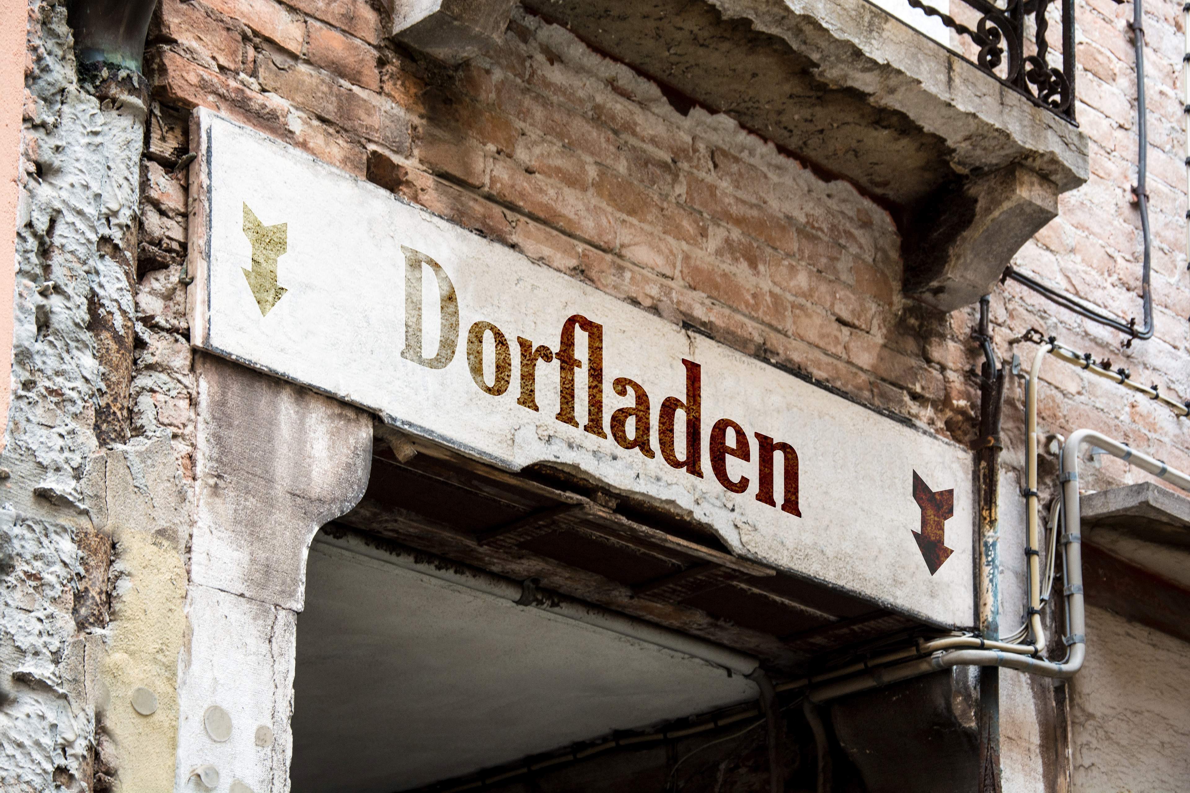 Zu sehen ist ein Schild mit der Aufschrift "Dorfladen", welches an einer alten Hauswand über einem Durchgang hängt. 