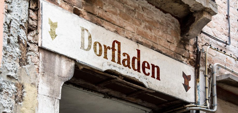 Zu sehen ist ein Schild mit der Aufschrift "Dorfladen", welches an einer alten Hauswand über einem Durchgang hängt. 