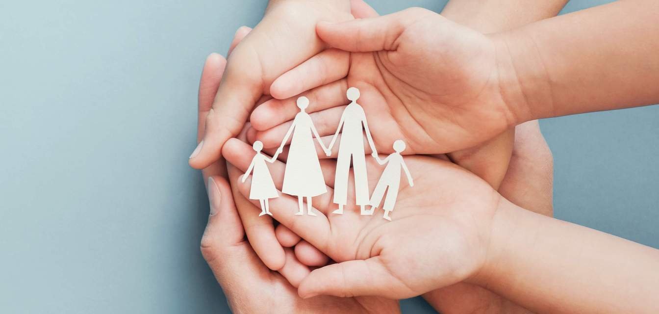 Vier übereinandergelegte Hände halten eine aus Papier ausgeschnittene Familie.