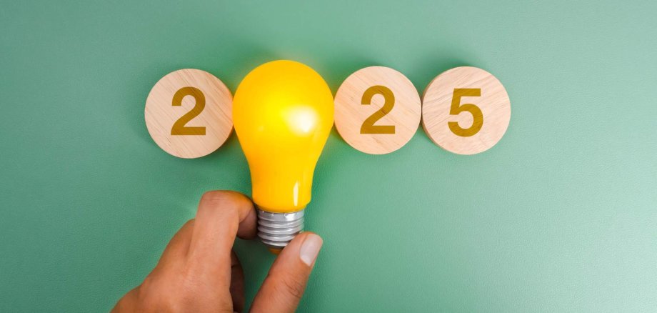 2025 Kalendernummer auf Holzblöcken mit einer leuchtend gelben Glühbirne, die in der Hand eines Mannes auf grünem Hintergrund gehalten wird.