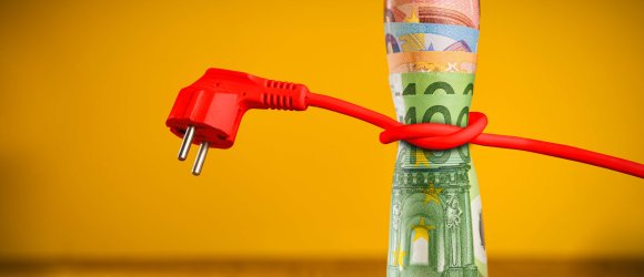 Um eine Rolle mit 100-Euro-, 50-Euro- und 20-Euro-Scheinen ist ein rotes Stromkabel gewickelt. Der Netzstecker vom Kabel links neben der Geldrolle zu sehen ist. Das Kabel stranguliert die Geldscheine.