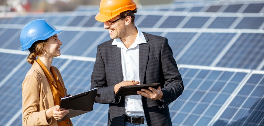 Ein Mann und eine Frau stehen mit Bauhelmen und Tablets vor Photovoltaik-Anlagen