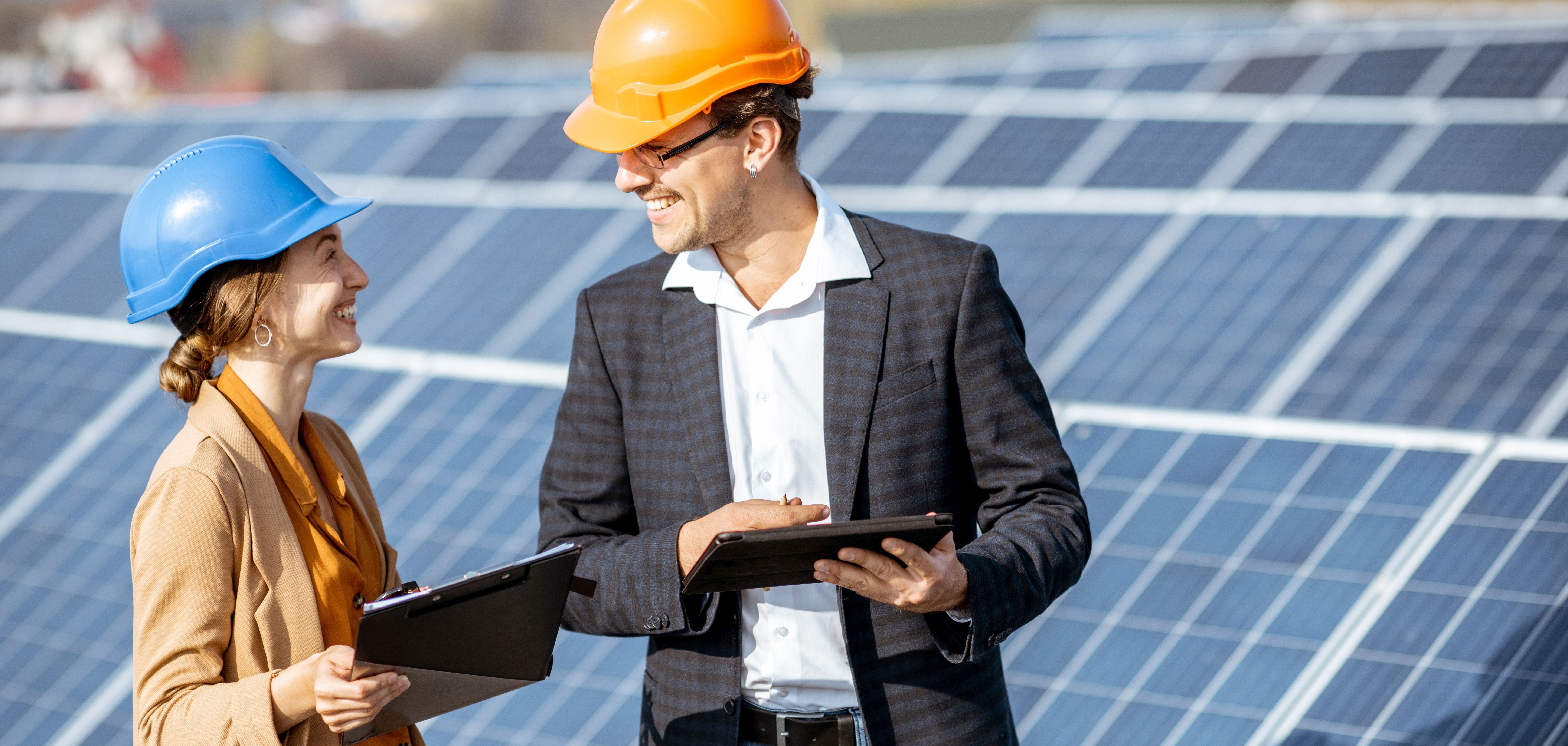 Ein Mann und eine Frau stehen mit Bauhelmen und Tablets vor Photovoltaik-Anlagen