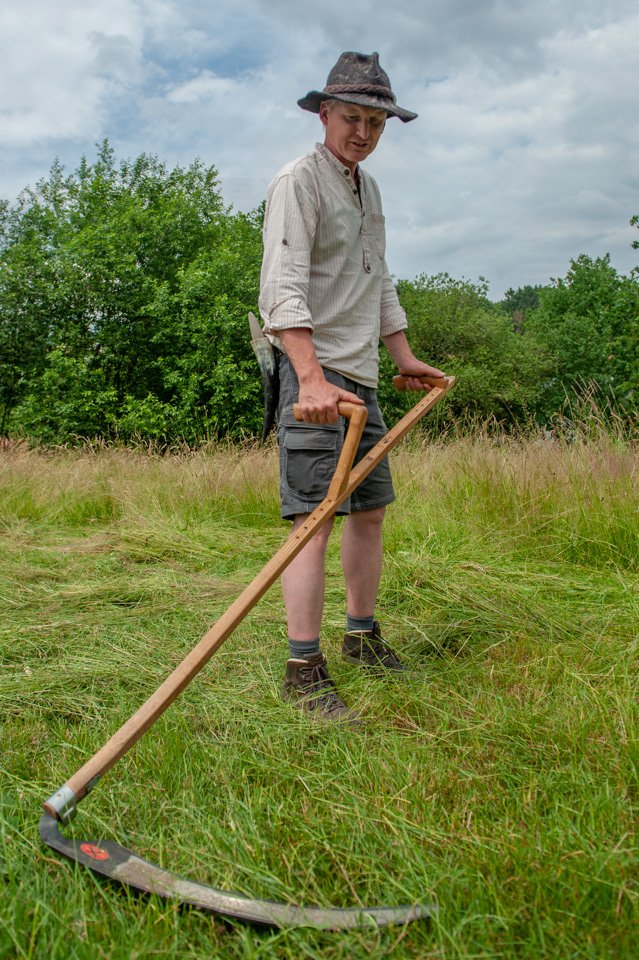 Sensen Kurs Man sieht im Vordergrund eine Sense, mit der Wiese gemäht wird. Im Hintergrund sieht man den Mann, der die Sense hält.