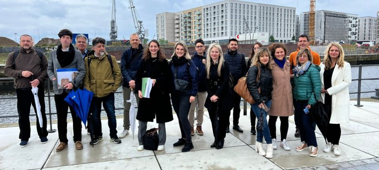 16 Personen (Männer und Frauen) stehen vor der Hamburger Hafen Skyline und lächeln in die Kamera.