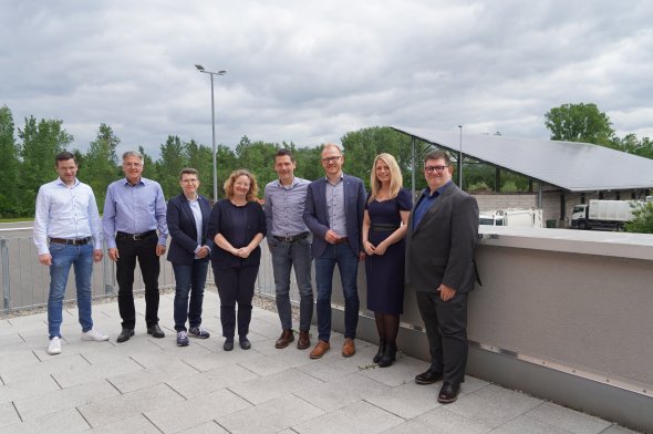 Acht Personen stehen auf einer Dachterrasse und lächeln in die Kamera. Im Hintergrund sieht man Müllfahrzeuge sowie eine große auf einem Dach befestigte PV-Anlage.