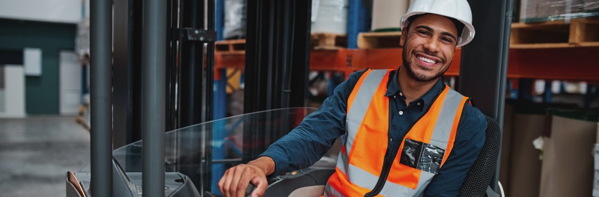 Portrait of happy young african forklift manager wearing safety vest and white hardhat transporting goods from one shelf to another while looking in camera in warehouse