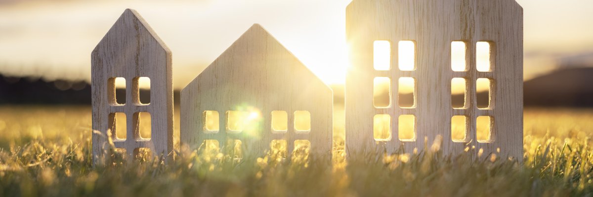 Umrisse dreie Häuser aus Holz stehen auf einer grünen Wiese, im Hintergrund geht die Sonne unter