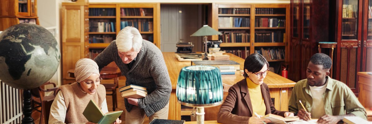 Diverse Menschen in der Bibliothek lernen an einem Holztisch. Im Hintergrund sind volle Bücherregale