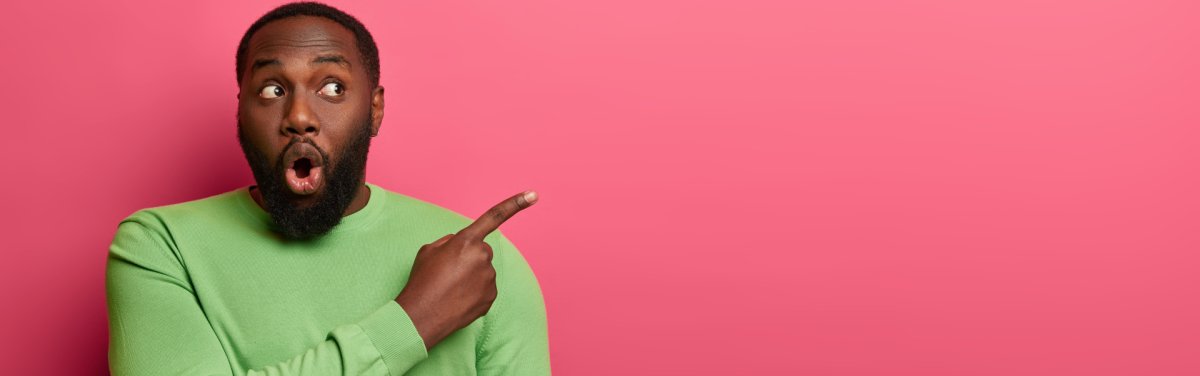 Shocked emotive bearded man with black skin, wears bright green sweater, points at empty space, surprised by unexpected relevation, demonstrates place on pink wall for your promotional content