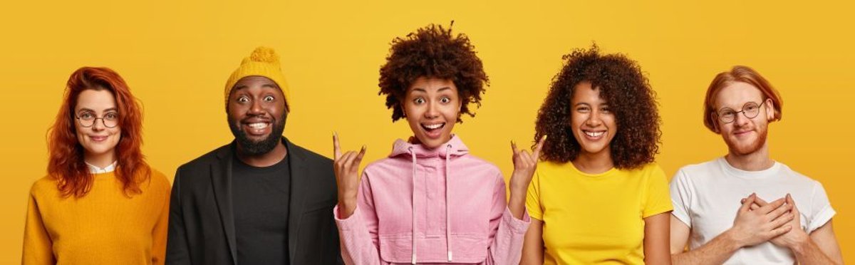 Positive mixed race friends pose together, stand next to each other. African American woman shows rock n roll gesture, ginger man keeps hands on heart, expresses gratitude, smile positively.