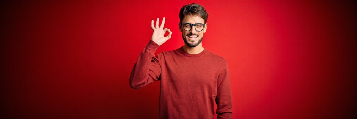 Young handsome man with beard wearing glasses and sweater standing over red background smiling positive doing ok sign with hand and fingers. Successful expression.