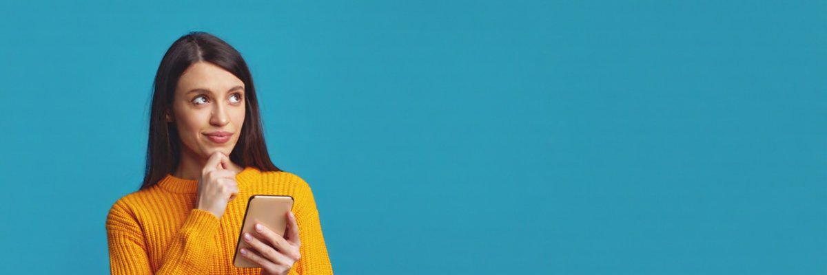 Pretty girl has holds chin thinks about mobile online offer, looks away contemplates about something, isolated over blue background with empty space  