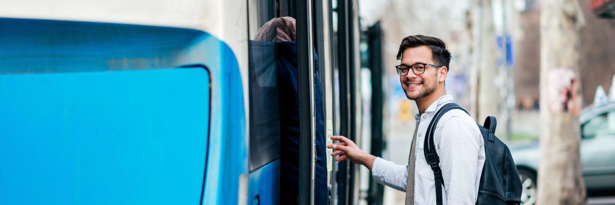 Ein junger dunkelhaariger Mann mit Brille steigt lächelnd in einen blauen Bus.