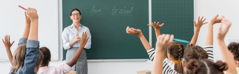 back view of multicultural pupils with hands in air, and teacher standing at chalkboard with back to school inscription, horizontal image Eine Unterrichtssituation mit Blick in Richtung Tafel.