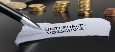 Auf einer schwarzen Fläche liegt ein karierter Zettel mit der Aufschrift §Unterhaltsvorschuss". Im Hintergrund sind mehrere Stapel Münzen und ein Portemonnaie zu sehen. 