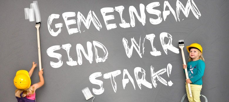 drei Kinder malen mit weißer Farbe den Schriftzug "gemeinsam sind wir stark" auf eine graue Wand.