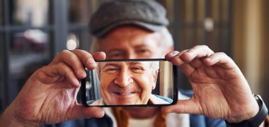 Ein alter Mann hält seinen Smartphonebildschirm mit beiden Händen in die Kamera, dabei verdeckt er sein Gesicht mit dem Handybildschirm zur Hälfte. Man sieht sein lächelndes Gesicht dafür auf dem Smartphonebildschirm.