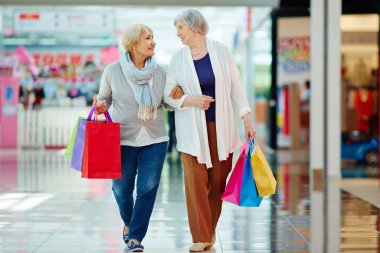 Zwei Seniorinnen laufen beieinander eingehakt durch ein Einkaufcenter und haben bunte Tüten in der freien Hand. 